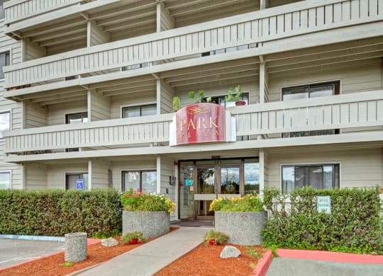 Facade of a beige building with a "Park Plaza" sign, featuring balconies and entrance surrounded by greenery and colorful flower planters.