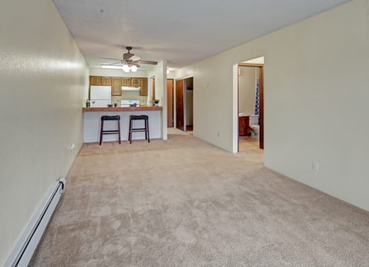 Long carpeted living room here at Park Plaza Apartments leading to an open kitchen with wooden cabinets and a ceiling fan. Two bar stools at a counter. Neutral tones, calming ambiance.