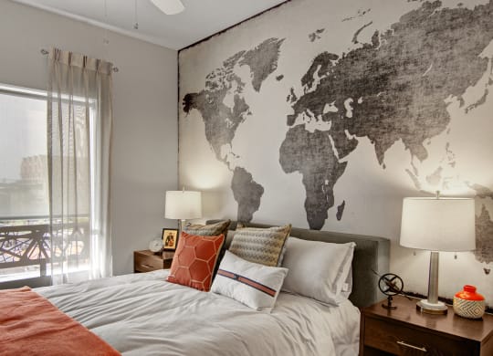 Cozy bedroom here at Proxy 333 Apartments with a world map mural behind the bed. Neutral tones, accented by orange pillows and throw on the bed, create a warm and inviting atmosphere.