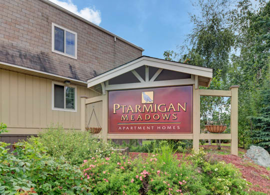 Sign reading "Ptarmigan Meadows Apartment Homes," surrounded by lush greenery and flowers, sits in front of a beige building under a partly cloudy sky.