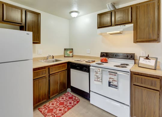 Compact kitchen here at Ptarmigan Meadows with wood cabinets, white appliances, and a red kettle on the stove. A red patterned rug adds warmth. Decor includes coffee-themed towels.
