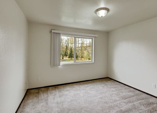 Small, empty bedroom here at Ptarmigan Meadows with beige walls and carpet. A single window with vertical blinds shows a view of trees outside. Ceiling light fixture adds a calm tone.