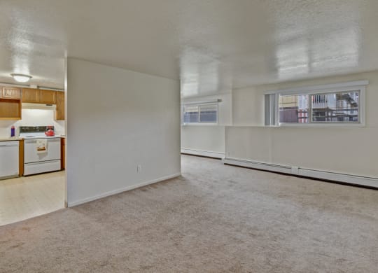 Spacious unfurnished room here at Ptarmigan Meadows with beige carpet and white walls. Adjacent kitchen has wooden cabinets, stove, and dishwasher. Large window offers natural light.