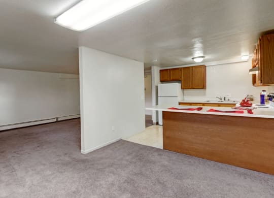 Open-plan room here at Ptarmigan Meadows with beige walls, carpeted living area, and a kitchen featuring wooden cabinets and a white fridge. A cozy, neutral space.