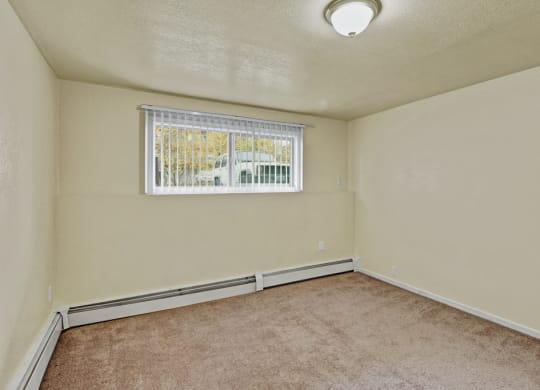 Empty beige bedroom here at Ptarmigan Meadows with brown carpet, a ceiling light, and a window with blinds overlooking a parked car. The space feels calm and neutral.