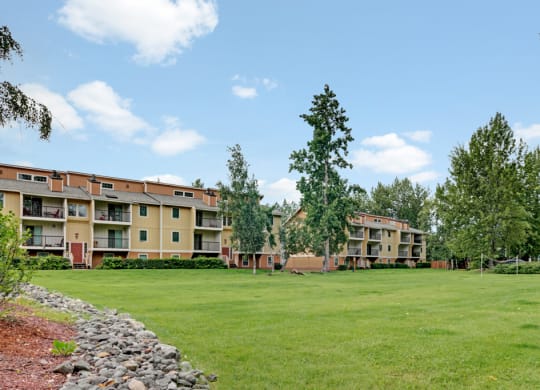 Ptarmigan Meadows Apartment complex with yellow-brown buildings, surrounded by trees and an expansive grassy area. Bright, sunny day with a few clouds in the sky.