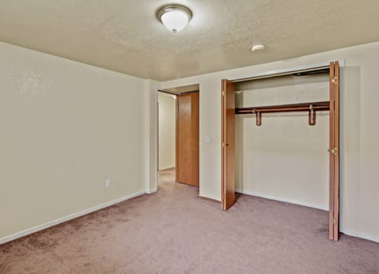 An empty bedroom here at Ptarmigan Meadows with beige walls and a brown carpet. Open closet with wooden doors and a hanging rod. Ceiling light illuminates the space, creating a simple, neutral tone.