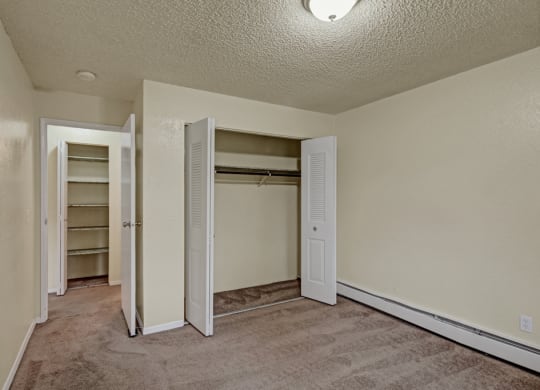 Small empty bedroom here at Ptarmigan Meadows with beige walls and carpet, featuring an open closet with louvered doors and a doorway leading to a shelf-lined closet, lit by a ceiling light.