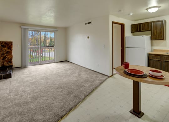 Cozy apartment interior here at Ptarmigan Meadows with beige carpet, sliding glass doors, small brick fireplace, and compact kitchen. A round table holds red dinnerware, creating a warm, inviting atmosphere.