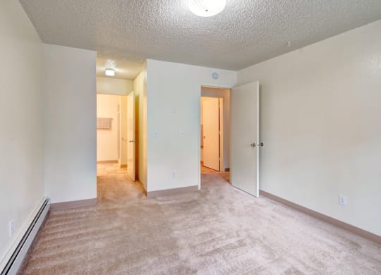 Empty bedroom here at Quiet Creek Apartments with beige carpet and off-white walls. An open door reveals a lit hallway. The room is minimalistic and calm, evoking a neutral tone.
