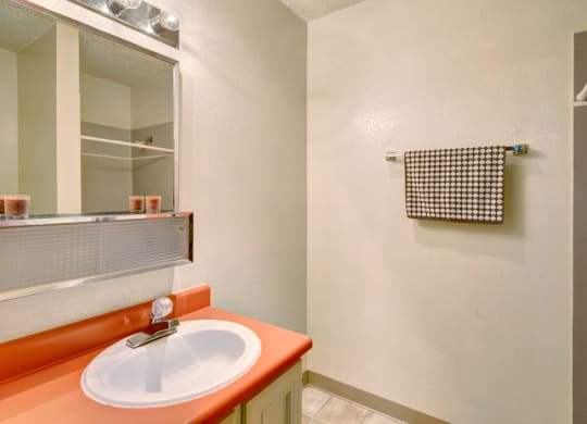 A small bathroom here at Quiet Creek Apartments with a coral countertop sink, a lit mirror above, and a patterned towel on a rack. Neutral tones create a clean, simple feel.