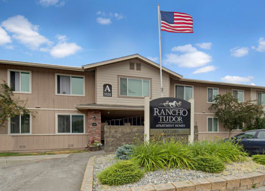 Two-story apartment building with beige siding, marked "A 3531." A U.S. flag waves above the "Rancho Tudor Apartment Homes" sign amidst landscaping.