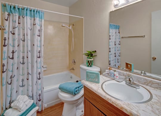 Bathroom here at Rancho Tudor Apartments with nautical-themed shower curtain featuring anchors, white bath, and sink with marble countertop. Towel, plant, and decor add a beach vibe.