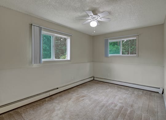 Empty bedroom here at Rancho Tudor Apartments with beige carpet and walls, featuring two large windows with vertical blinds. A ceiling fan with a light fixture hangs in the center.