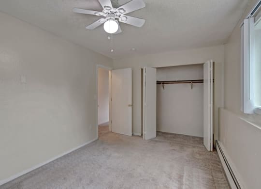 Empty bedroom here at Rancho Tudor Apartments with beige walls and carpet, featuring an open closet, a ceiling fan with light, and a window on the right letting in soft natural light.