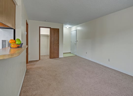 Empty living room here at Rancho Tudor Apartments with beige carpet and white walls, featuring a wooden door to a closet, a white entrance door, and a bar counter with a fruit bowl.