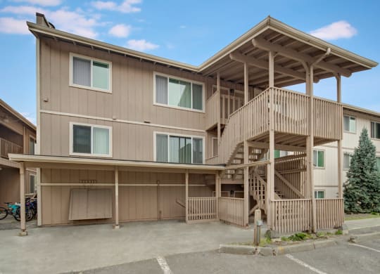 Three-story beige Rancho Tudor Apartment building with prominent wooden staircase and railings, set against a blue sky. Nearby are parked bicycles and evergreen trees.