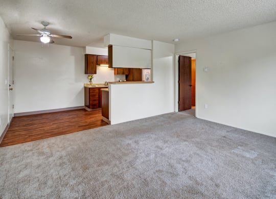 Spacious empty living room here at Rancho Tudor Apartments with beige carpet, white walls, and a small kitchen area with wooden cabinets. Ceiling fan and open doorway add a welcoming feel.