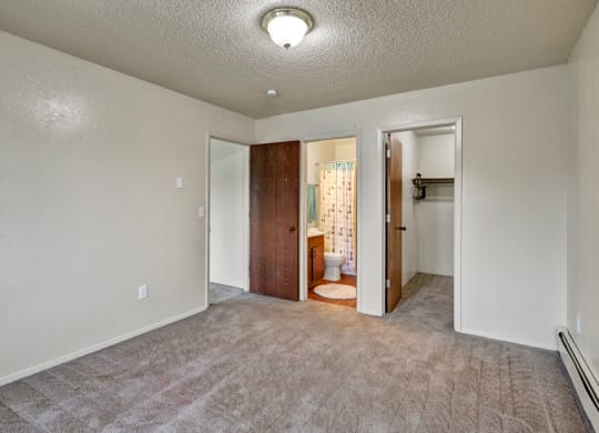 Empty carpeted room here at Rancho Tudor Apartments with beige walls, featuring two wooden doors. One door leads to a bathroom with a shower curtain; the other to a closet. A ceiling light softly illuminates the space.