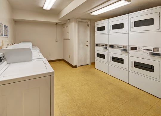 Laundry room here at Rancho Tudor Apartments with white machines and beige floor. Six stacked dryers are on the right, and five washing machines are on the left. Bright fluorescent lights illuminate the room.