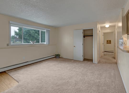 Empty beige living room here at Rancho Tudor Apartments with a large window showing greenery outside. The room features carpet, an open closet with a door, and a hallway with dim lighting. Calm and neutral ambiance.