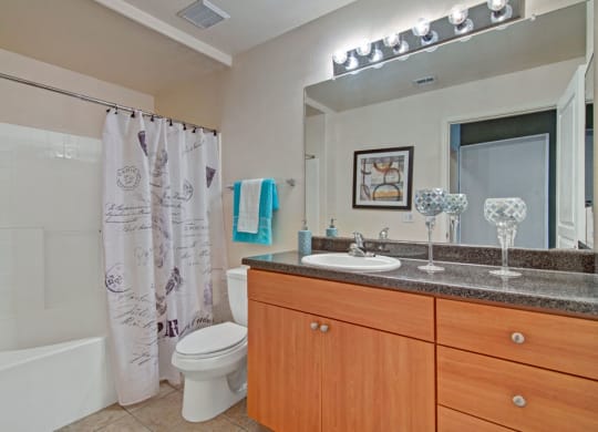 Modern bathroom here at Rio Paradiso Apartments with wooden vanity and granite countertop. A large mirror with bright lights, decorative candles, and a patterned shower curtain add elegance.