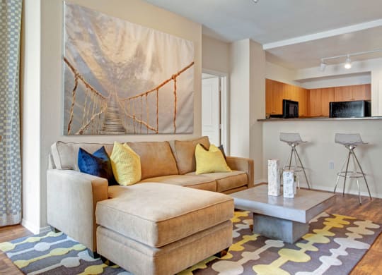 Cozy living room here at Rio Paradiso Apartments with beige sectional sofa, colorful pillows, and abstract rug. Features a large bridge photo on the wall and sleek bar stools by the kitchen.