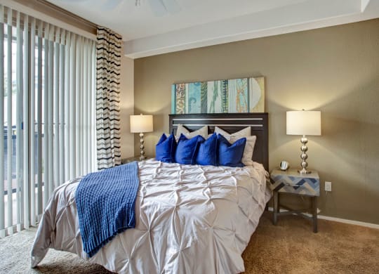 A cozy bedroom features a neatly made bed with a cream comforter and blue pillows here at Rio Paradiso Apartments. Two lamps on wooden nightstands illuminate the space. Vertical blinds cover a large window, creating a serene atmosphere.