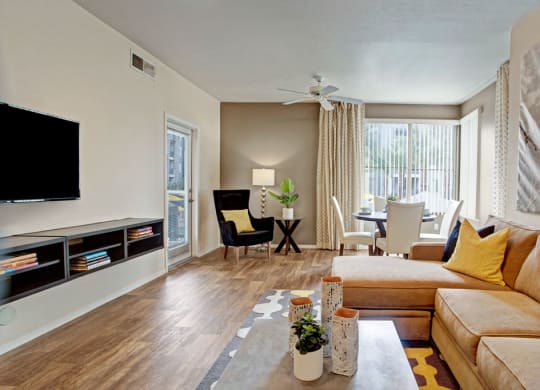 A modern living room here at Rio Paradiso Apartments with a beige sectional sofa, yellow cushions, and a geometric rug. A wall-mounted TV is opposite, with a dining area by a large window. Bright and inviting atmosphere.