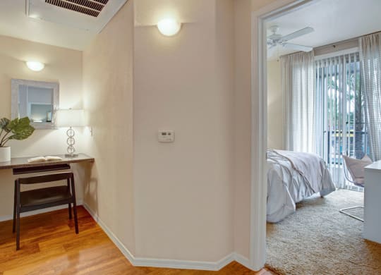 Cozy interior view here at Rio Paradiso Apartments with a small desk nook on the left featuring a mirror, potted plant, and lamp. To the right, a bright bedroom with sheer curtains and a ceiling fan.