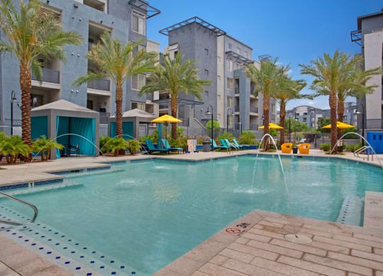 Rio Paradiso Apartment complex pool area with palm trees and blue-gray buildings. Features lounge chairs, yellow umbrellas, cabanas, and water fountains, creating a relaxing atmosphere.