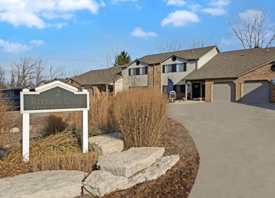 Sign reading "River's Cove Apartment Homes" near a pathway leading to brick and siding apartments with garages under a clear blue sky.