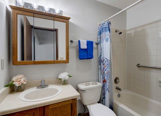 Small, tidy bathroom here at Russian Jack Apartments with a white sink, wooden cabinet, and mirror. A flower arrangement and blue towels add color. Shower has a patterned curtain.