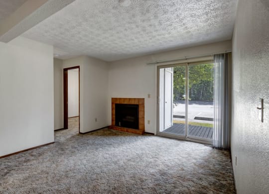 Empty living room here at Russian Jack Apartments with textured ceiling, beige carpet, and neutral walls. Features a brick fireplace, sliding glass door with vertical blinds, and natural light. Atmosphere feels calm and open.