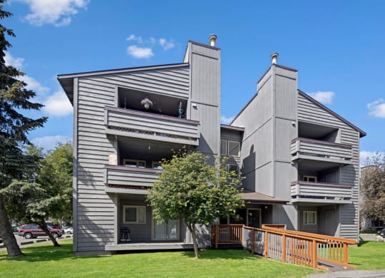 Gray three-story Russian Jack Apartment building with balconies, set against a backdrop of blue sky and trees. A wooden ramp leads to the entrance. Calm atmosphere.