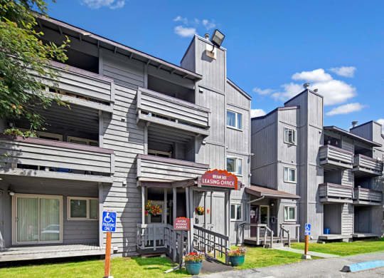 Gray, multi-story Russian Jack Apartment building with balconies under a blue sky. A leasing office sign is visible. Accessible parking signs are in front.