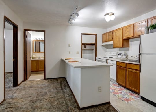 Compact kitchen here at Russian Jack Apartments with wooden cabinets, white appliances, and a floral rug. A long countertop extends into a hallway showing a lit bathroom mirror. Cozy feel.