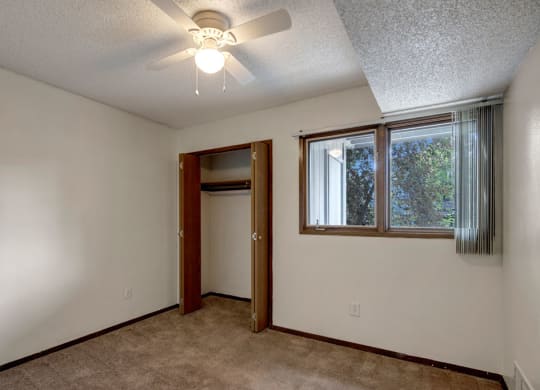 Small, empty bedroom here at Russian Jack Apartments with beige carpet, white walls, and a ceiling fan. Open closet with wooden doors and a window on the right with vertical blinds.