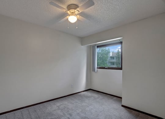 Small, empty room here at Russian Jack Apartments with light gray walls, a ceiling fan, and a window showing trees outside. Carpeted floor adds a cozy feel. Bright, serene atmosphere.