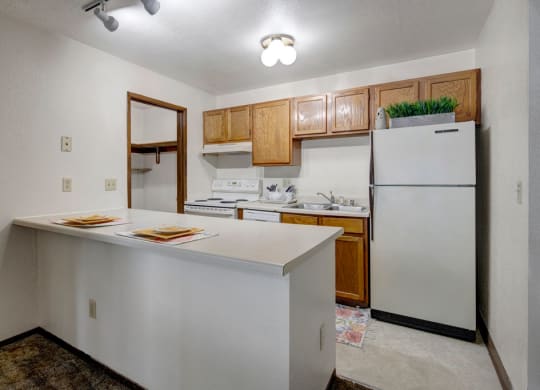 Small, cozy kitchen here at Russian Jack Apartments with overhead lighting, featuring wooden cabinets, a white fridge, stove, and sink. Simple counter with two place settings.