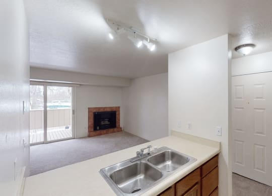 Small, empty living room here at Russian Jack Apartments with beige walls and carpet. Features a fireplace, sliding glass door to a balcony, and a kitchen view with a double sink. Neutral lighting.