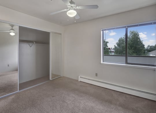 Empty bedroom here at Silver Ridge Apartments with beige carpet and white walls. Large mirrored closet doors reflect a ceiling fan. Bright window view reveals green trees and blue sky.