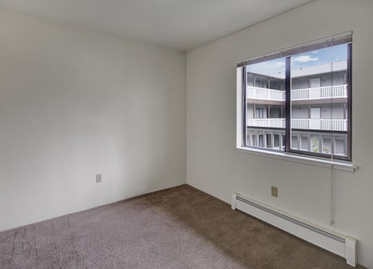 Empty bedroom here at Silver Ridge Apartments with beige carpet, white walls, and a window showing an apartment building courtyard. The light creates a calm, neutral tone.