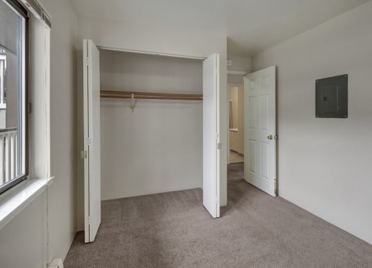 Empty bedroom here at Silver Ridge Apartments with beige carpet, open closet with wooden rod, white walls, and doors leading to another area. A window on the left brings in light.