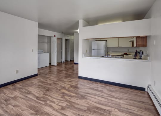 Spacious, empty living room here at SIlver Ridge Apartments with light wood flooring and white walls. Open kitchen with a visible fridge and countertop, providing a clean, minimalist feel.