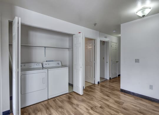 Bright laundry area here at Silver Ridge Apartments with white washer and dryer set against a light wall. Wood flooring leads to open doors revealing more rooms, under soft overhead lighting.