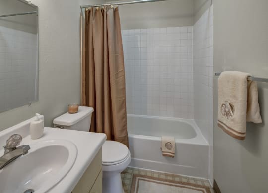A small bathroom here at Silver Ridge Apartments with white sink and toilet, beige shower curtain, and matching towels with shell motifs. A candle adds a warm, inviting touch.