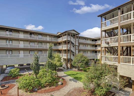 Four-story Silver Ridge Apartment complex with beige and brown tones, featuring outdoor staircases and white railings. A landscaped courtyard has trees, shrubs, and a gravel path under a sunny blue sky.