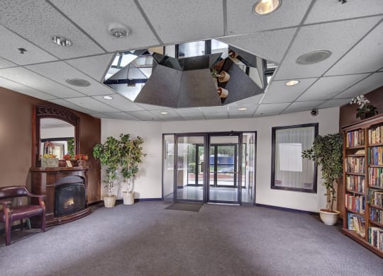 Lobby here at Silver Ridge Apartments with a cozy and inviting feel, featuring a dark wooden fireplace with a mirror above it, two potted plants, a bookshelf full of books, and seating.