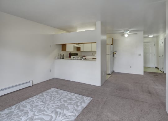 Spacious living room here at Silver Ridge Apartments with beige carpet and patterned rug. Open view of a white kitchen with appliances. Ceiling fan and soft lighting create a cozy atmosphere.